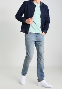 Marineblauwe bomberjack over een lichtgroen T-shirt, gecombineerd met lichtblauwe spijkerbroek en witte sneakers. Casual pasvorm met een gladde textuur.