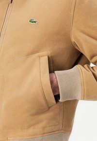 Veste brune avec une texture lisse, dotée d'une fermeture éclair à l'avant et de poignets côtelés ; inclut un petit logo vert brodé sur la poitrine.