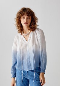 Blusa leggera blu e bianca con gradiente, scollo a V, maniche lunghe e dettagli con cordoncino. Abbinata a pantaloni con motivo floreale blu.
