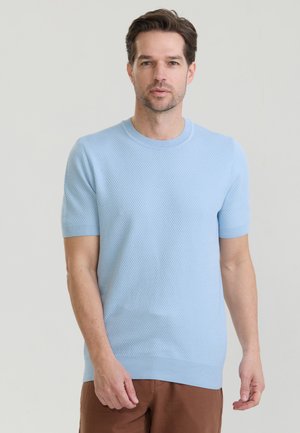 Mężczyzna o krótkich brązowych włosach, ubrany w jasno niebieski teksturowany sweter z krótkim rękawem i brązowe spodnie, stojący na jednolitym tle.