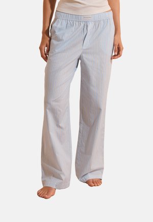 Persona che indossa pantaloni del pigiama larghi a righe azzurre e bianche, in piedi a piedi nudi con le mani rilassate lungo i fianchi.