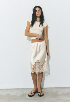 Donna con lunghi capelli scuri che indossa un crop top crema e una gonna rifinita con pizzo, in piedi contro un muro bianco con sandali neri.