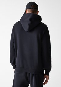 Felpa con cappuccio blu navy dal texture morbido, con polsini e orlo a costine. Presenta un ampio cappuccio strutturato e una silhouette rilassata.