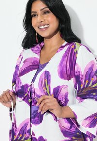 Blomster cardigan med store lilla liljeprint på hvid baggrund, med båndlukning og klokkeærmer, lagdelt over en lilla top.