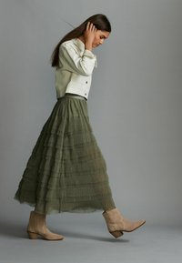 Gonna maxi verde a balze con pieghe orizzontali, abbinata a una giacca di jeans bianca e stivaletti beige alla caviglia. Texture lisce e design a strati.