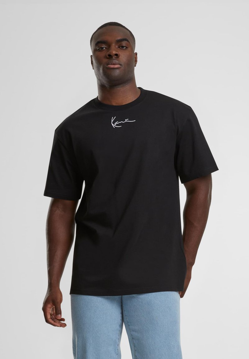 Homme portant un T-shirt noir avec un logo blanc en écriture sur la poitrine et un jean bleu clair, debout devant un fond gris uni.