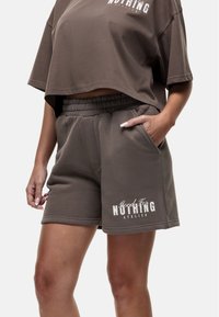 Bruine katoenen shorts met een elastische tailleband en zijzakken. Bevat witte tekst met de tekst "Goods For Nothing Atelier" op de zoom.