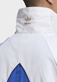 White athletic jacket with a high collar, featuring a blue triangular accent and gold embroidered Adidas logo on the back. Smooth texture.
