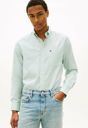 Mężczyzna w jasnozielonej koszuli na guziki z logo na kieszeni piersiowej i jasnoniebieskich dżinsach, stojący z jedną ręką w kieszeni.