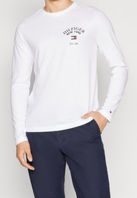 Weißes Langarm-T-Shirt aus Baumwolle. Mit einem schwarzen Logo mit dem Text "HILFIGER NEW YORK" und roten sowie blauen Akzenten. Schlichtes Design.