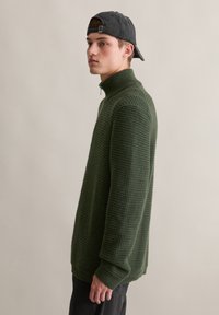 Grüner gestrickter Pullover mit hohem Kragen, der ein strukturiertes Muster aufweist, lange Ärmel hat und eine lässige Passform bietet, kombiniert mit einer Mütze.