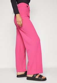 Pantalon rose à jambes larges avec une texture lisse, doté d'une taille élastique et de poches latérales, associé à des sandales plates noires.