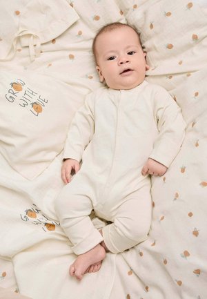 Baby in beigem Strampler liegt auf gemusterten Bettlaken mit kleinen orangefarbenen Fruchtmotiven und einer Stofftasche mit der Aufschrift "GROW WITH LOVE."