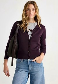 Violetti neulottu neuletakki napillisen suljettavan, jossa on resorireunus ja resoriset hihansuut, yhdistetty harmaaseen t-paitaan ja vaaleansiniin farkkuihin.