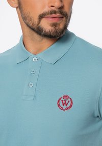 Jasnoniebieska koszulka polo z teksturowanej tkaniny, z trzema guzikami z przodu oraz czerwonym haftowanym logo z wzorem laurowym na piersi.