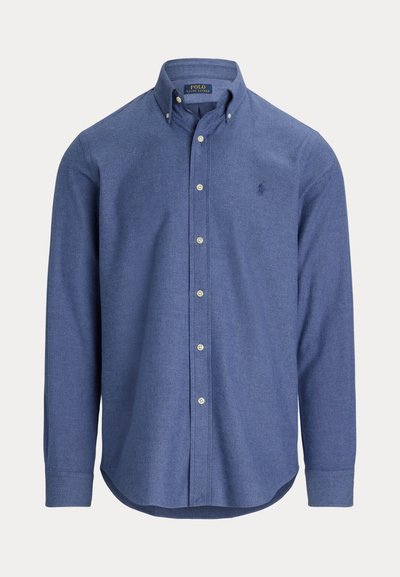 Sininen nappikauluspaita, joka on valmistettu tekstuurikankaasta ja jossa on terävä kaulus, pitkät hihat ja pieni logo vasemmassa rinnassa.