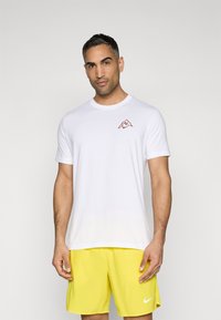 Hvit T-skjorte med kort erm som har en rød fjell-logo på venstre bryst; glatt stoff; kombinert med lyse gule shorts.
