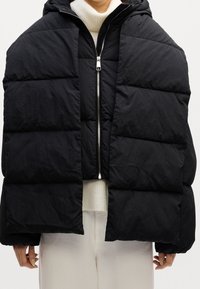 Personne portant une doudoune noire oversize avec un matelassage horizontal, une fermeture éclair argentée et une capuche sur un pull blanc et un pantalon clair.