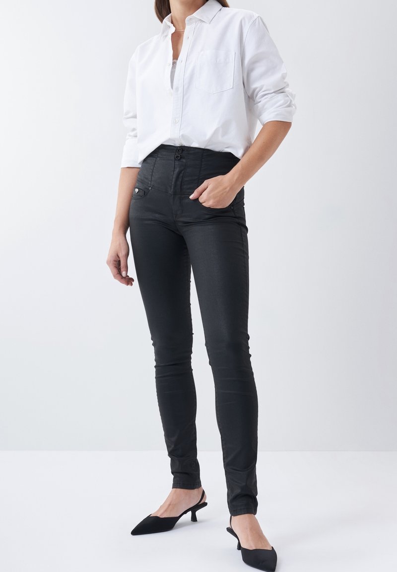 Femme portant une chemise blanche boutonnée, un pantalon skinny noir taille haute, et des chaussures slingback noires à petit talon, debout devant un fond blanc uni.