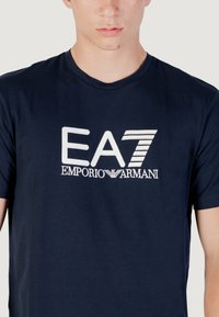 Camiseta de algodón azul marino con un logo bordado en blanco "EA7 EMPORIO ARMANI" en el pecho, manga corta y cuello redondo clásico.
