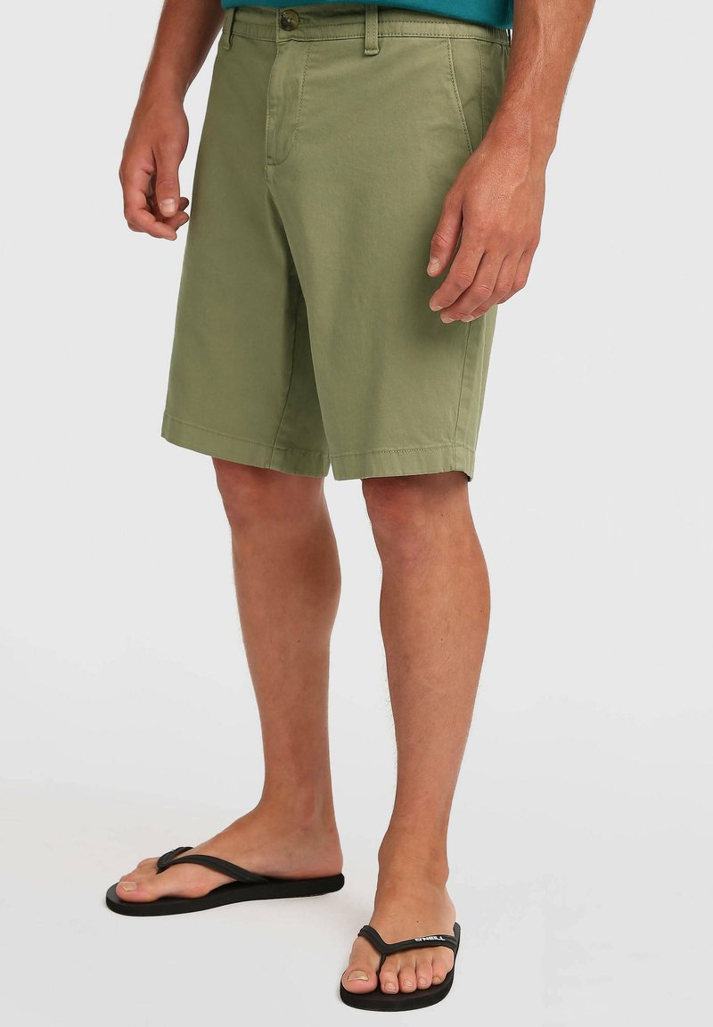 Pantalones cortos de algodón verde oliva con corte recto, bolsillos delanteros y cierre con botón, combinados con chanclas negras. Diseño y textura simples.