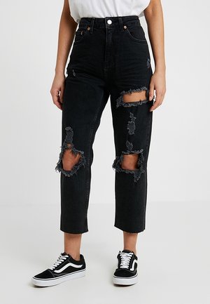 Jean noir taille haute déchiré avec de grandes déchirures aux cuisses et aux genoux, porté avec des baskets noires et blanches et un haut blanc.