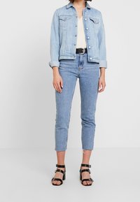 Light blue denim jacket with silver buttons and front pockets, paired with light blue high-waisted jeans, and black strappy heels.