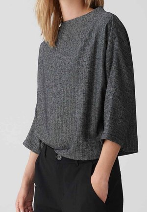 Frau trägt ein locker sitzendes graues Oberteil mit Fischgrätmuster und Dreiviertelärmeln sowie schwarze Hosen, eine Hand in der Tasche.