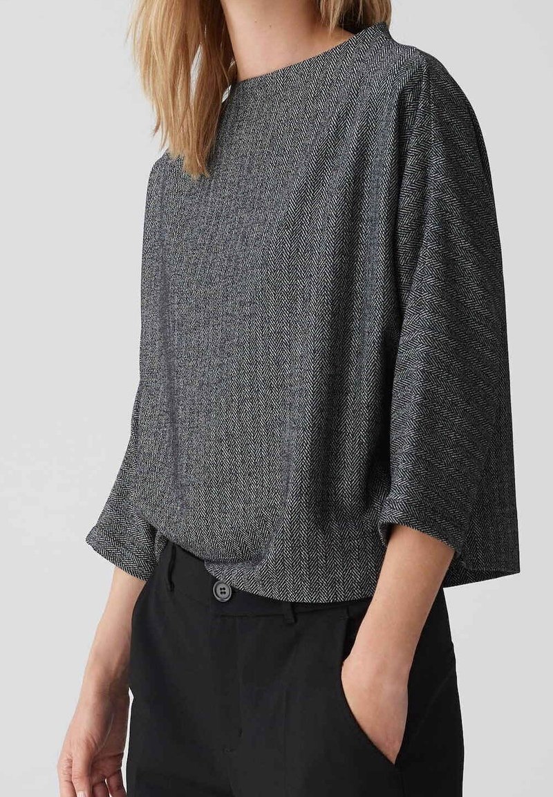 Femme portant un haut ample gris à motif chevrons avec des manches aux trois quarts et un pantalon noir, une main dans la poche.