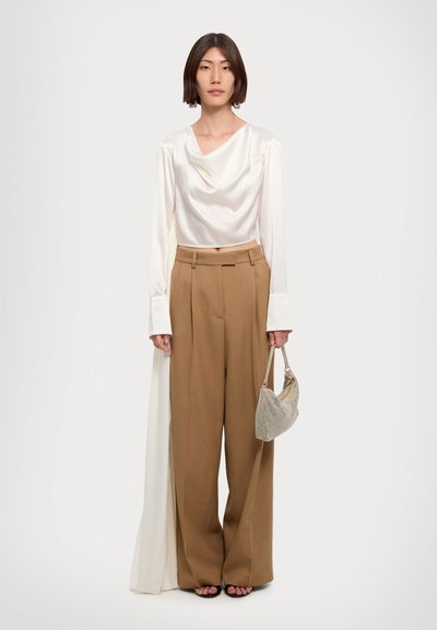 Beige ψηλόμεσες παντελόνες με χαλαρή γραμμή, σε συνδυασμό με ένα λευκό σατέν μπλούζα με στρογγυλή λαιμόκοψη. Το μοντέλο κρατά μια μικρή τσάντα με μεταλλική υφή.