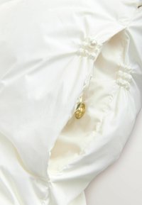Hvid satin-stof med tufted søm, små stofbetrukne knapper og en perledetalje nær en guld trykknap.