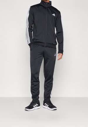 Mężczyzna w czarnym dresie Adidas z białymi paskami na rękawach i spodniach, w połączeniu z czarnymi butami sportowymi Adidas z białymi podeszwami.