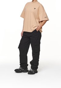 Bežové oversized tričko s malým černým logem, v kombinaci s černými cargo kalhotami s několika kapsami a černými sportovními botami s bílými tkaničkami.