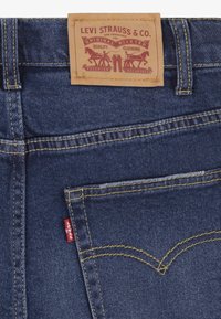 Dark denim jeans with a tan leather label, yellow stitching, and a red Levi's tab on the pocket. Features a classic design and riveted details.