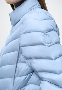 Ανοιχτό μπλε μπουφάν με ψηλό γιακά, με ραμμένη σχεδίαση και στρογγυλό λογότυπο στον ώμο. Ομαλή υφή.