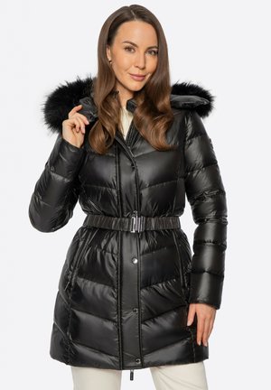 Femme portant un manteau matelassé noir ceinturé avec une capuche bordée de fourrure, tenant le bord de la capuche, debout devant un fond neutre.