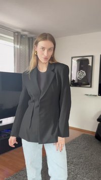 Blazer preto ajustado com lapelas bem definidas, ombros estruturados e corte reto. Usado sobre uma blusa cinza e combinado com jeans azul claro.