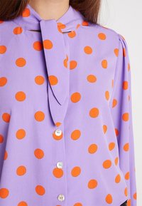 Blusa lavanda con grandi pois arancioni, caratterizzata da un dettaglio sul collo annodato, apertura con bottoni e tessuto morbido e leggero.