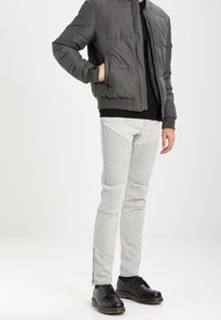 Harmaa toppatakki, jossa edessä vetoketju, musta neulepaita alla, yhdistetty vaaleanharmaisiin cargo-housuihin, joissa on teksturoidut yksityiskohdat ja vetoketjut, mustat kengät.