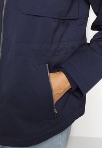 Nachtblaues Jackett mit Reißverschluss, aufgesetzten Taschen und schlanker Passform. Verfügt über einen gerippten Bund und eine glatte Stoffstruktur.