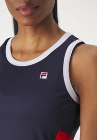 Navy tank top with white neckline, features a small red and blue Fila logo. Smooth texture with a fitted design. Red accent at the side.