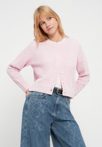 Ροζ πλεκτή ζακέτα με ριμπ ύφανση, στρογγυλή λαιμόκοψη και κουμπιά μπροστά. Συνδυασμένη με μπλε τζιν παντελόνι φαρδιάς γραμμής με μοτίβα.