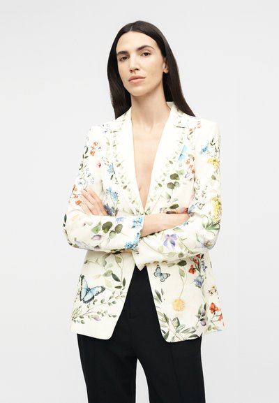 Γυναίκα με μακριά σκούρα μαλλιά, ντυμένη με λευκό σακάκι με πολύχρωμα λουλουδάτα και πεταλούδες σχέδια, στέκεται με σταυρωμένα τα χέρια μπροστά σε απλό φόντο.