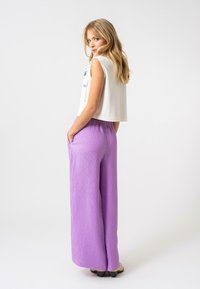 Avklippt vit topp med ficka, i kombination med flödande, texturerade lavendelfärgade vida byxor. Modellen står med händerna i fickorna, vänd något bakåt.
