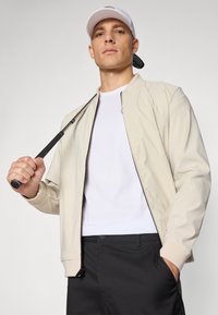 Beige bomber jacket with a smooth texture, black zipper detail, paired with a white t-shirt and black pants, holding a golf club.