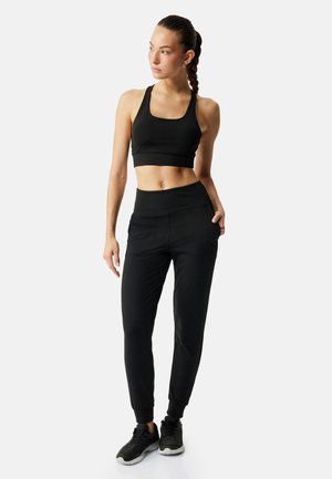 Kobieta z plecionymi włosami, ubrana w czarny sportowy stanik, spodnie dresowe i czarne sneakersy, stoi z jedną ręką w kieszeni, patrzy w lewo.