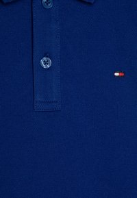 Mørkeblå polo skjorte laget av teksturert bomull, med en to-knapps knappestolpe og en liten brodert logo i rødt, hvitt og blått.
