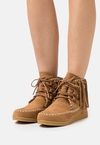 Béžové semišové kotníkové boty s třásněmi, bílým prošíváním a kulatými tkaničkami. Plochá gumová podrážka s texturovaným povrchem.