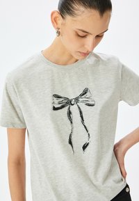 Grå bomulds t-shirt med en dekorativ sort snor-bue trykt med sølvfarvede rhinsten på forsiden. Rund halsudskæring, korte ærmer.