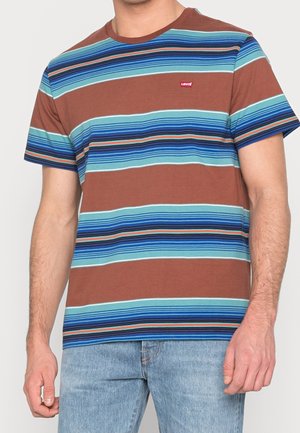 Mężczyzna ubrany w brązowy T-shirt z krótkim rękawem, z poziomymi niebieskimi i zielonkawymi paskami oraz jasnoniebieskie dżinsy.